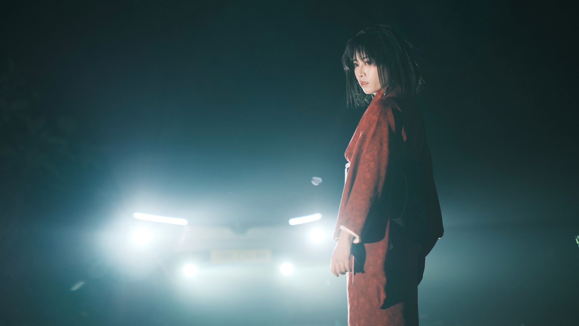 A woman in traditional clothing stands in front of car headlights at night, crea